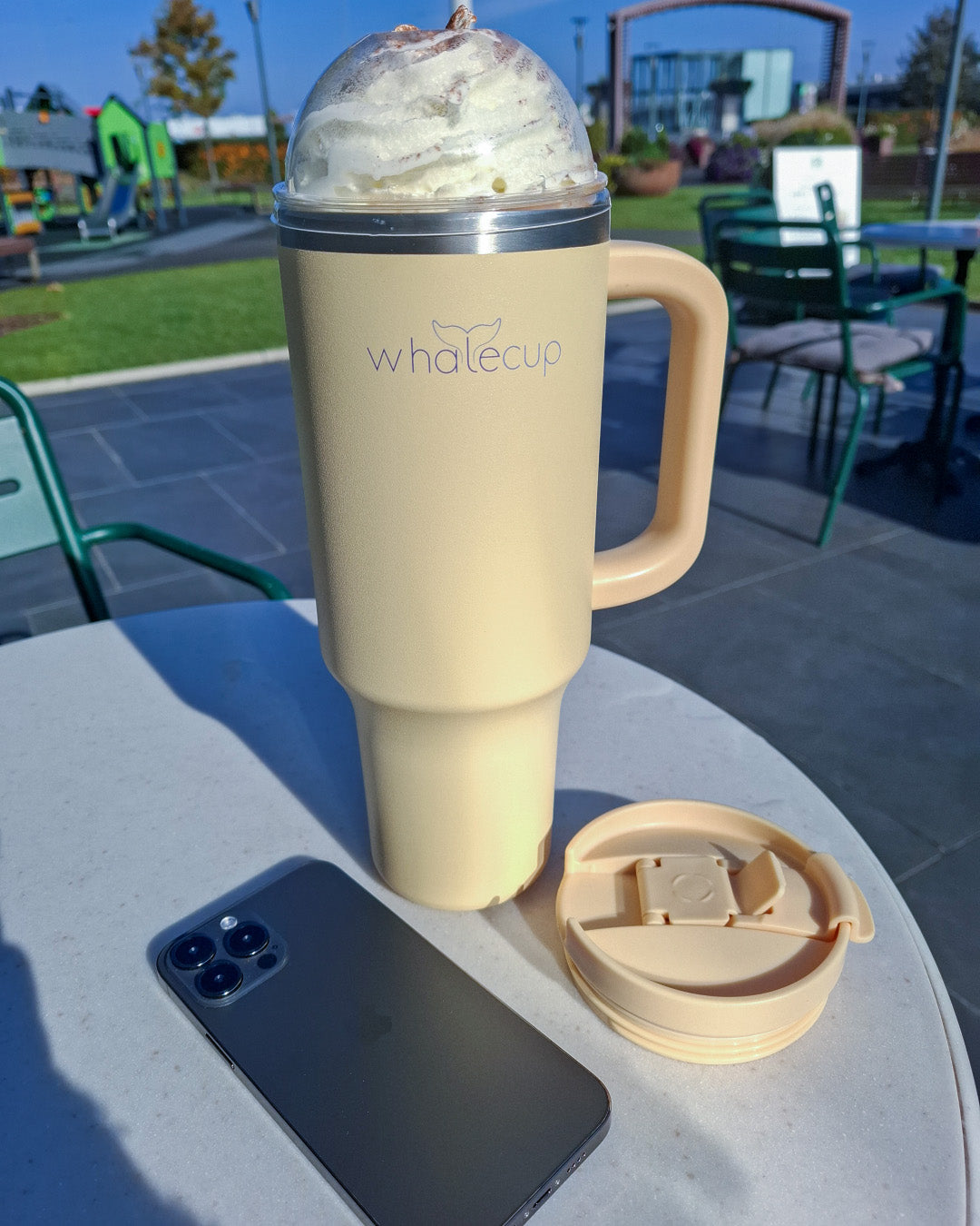 Whalecup cream tumbler filled with iced coffee and whipped cream, outdoor café table scene with phone and lid beside cup – stays cold and stylish